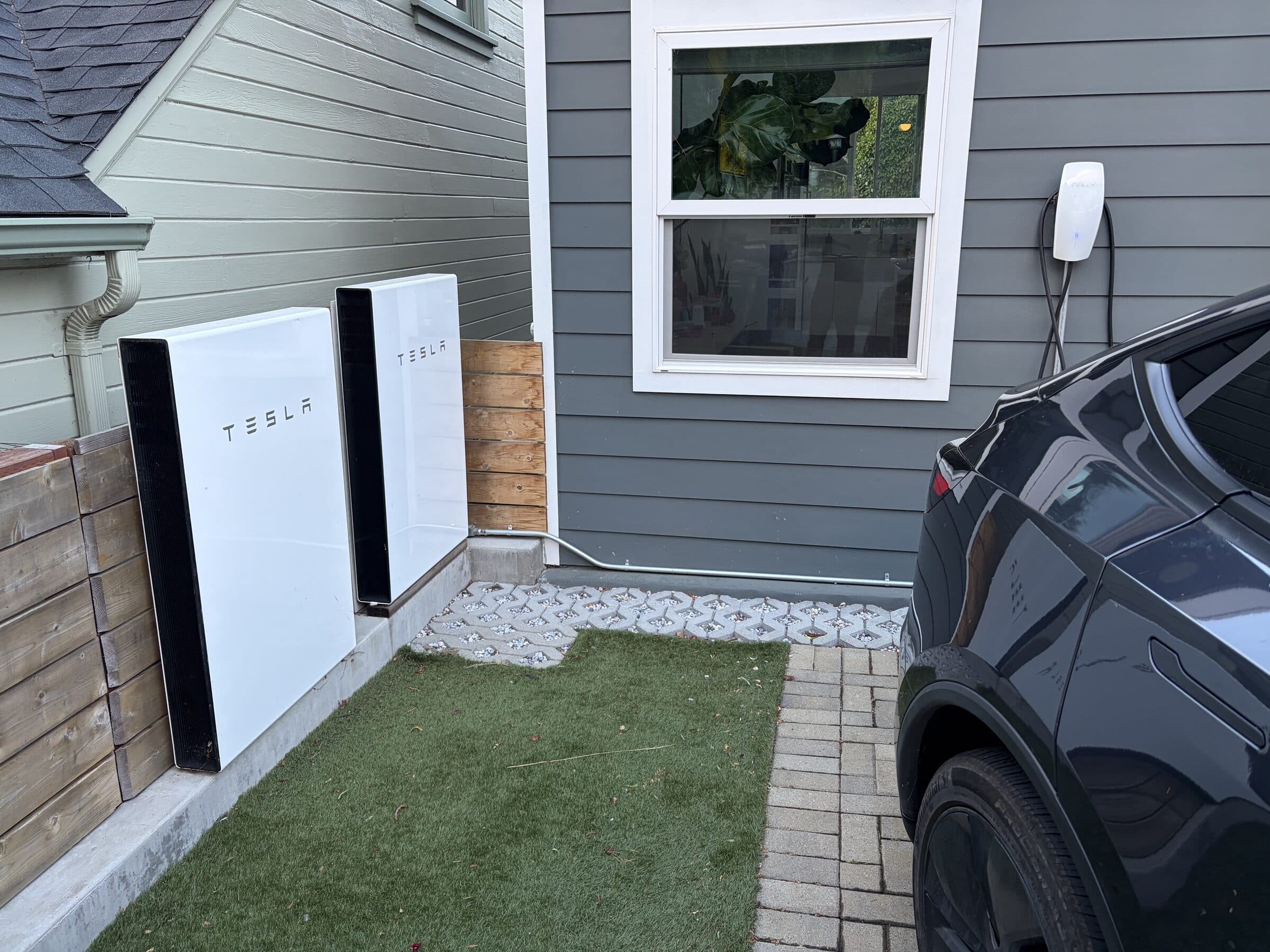 Two Tesla Powerwalls mounted on the exterior wall next to a Tesla wall connector and an EV charging in the driveway